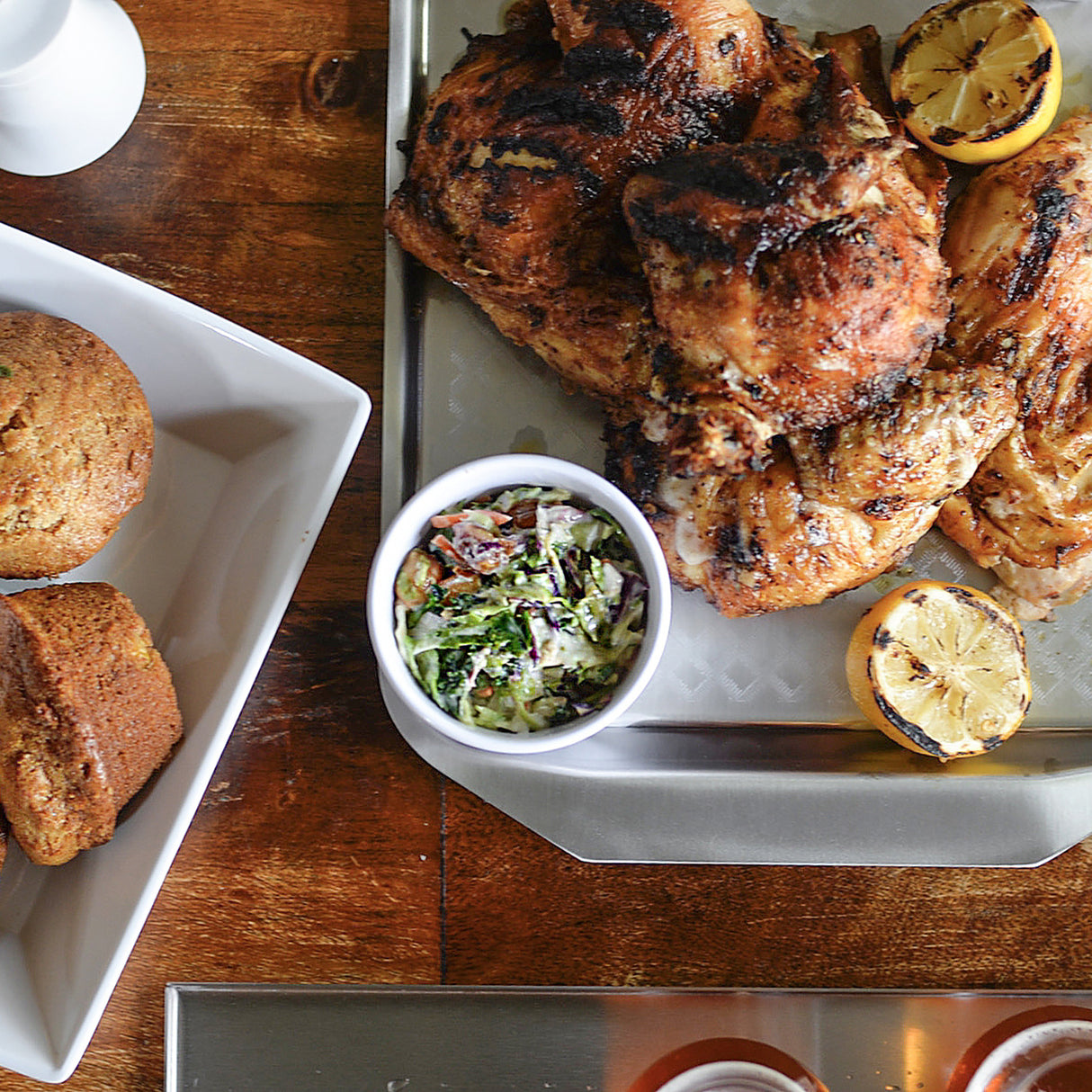 A metal tray with grilled chicken and charred lemon halves sits beside a soufflé cup filled with coleslaw. On the left, a white dish holds two golden brown muffins atop a wooden table, partially visible.