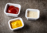 Three square melamine sauce cups from the Better Burger Collection sit on a gray, textured surface, each holding a different condiment—red ketchup, pale beige mayonnaise, and bright yellow mustard—in a neat triangular pattern.