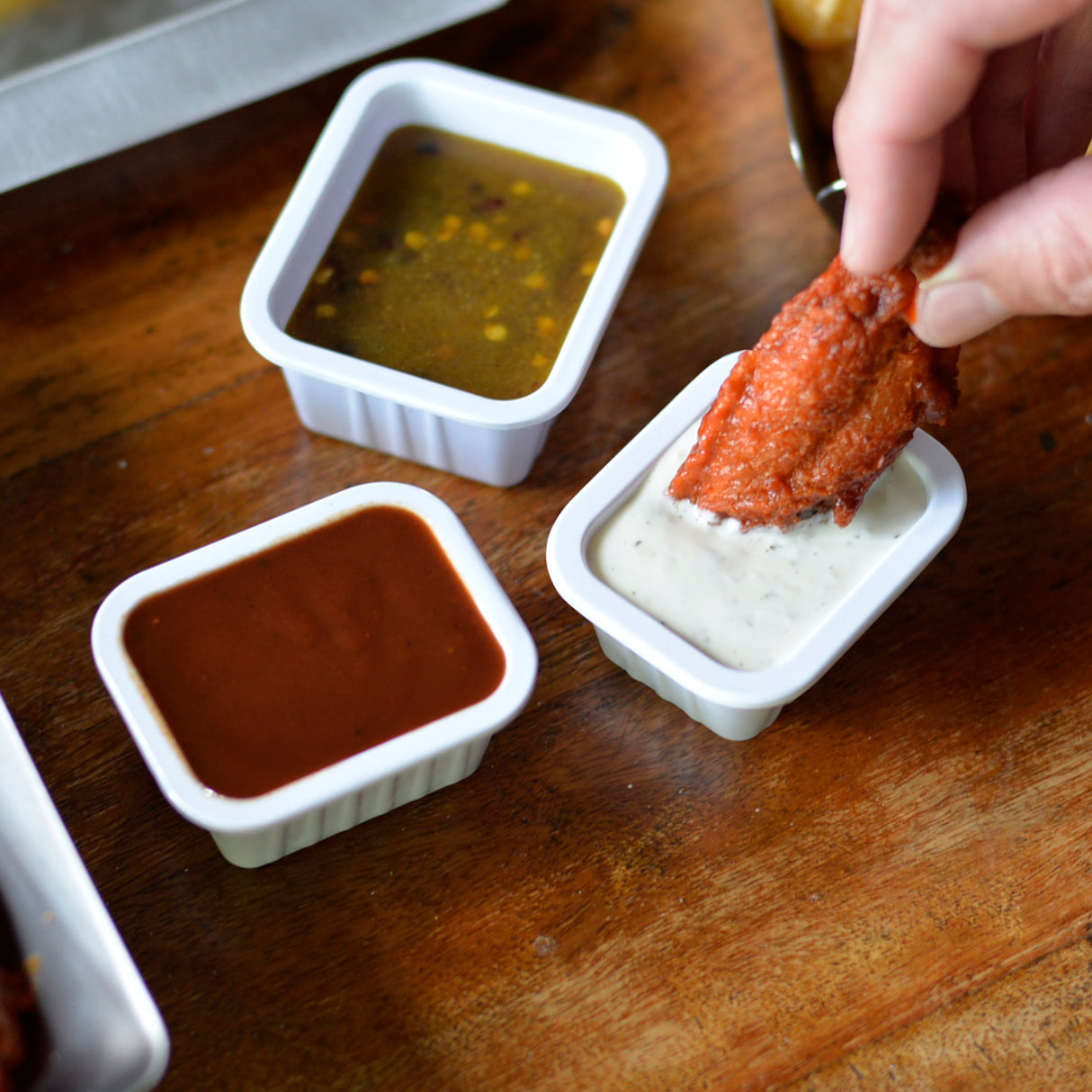 A hand dips a crispy, red-coated chicken wing into a small white melamine soufflé cup of creamy ranch dip. Next to it are two more soufflé cups filled with barbecue and vinaigrette sauces on a wooden table. All cups are dishwasher safe.