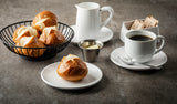 A breakfast scene with a cup of black coffee, a plate with a pretzel roll, a basket of more pretzel rolls, and a 3 oz stainless steel sauce cup filled with butter, plus a white milk pitcher and sugar packets container on a gray surface.