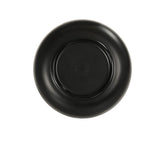 A black ceramic plate is shown from above, displaying its underside. The plate, reminiscent of an Oslo Bowl, has a glossy finish and a slightly raised base. There is a faint, circular maker’s mark with text and a logo in the center of the base.
