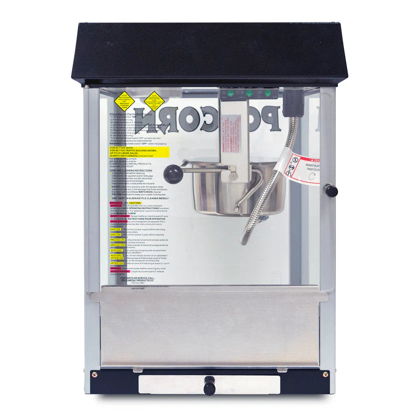 A stainless steel countertop popcorn machine with a glass front, black top, and silver base. Inside, a metal kettle is suspended, and safety warning labels are visible on the glass. POPCORN appears in large letters on the back wall.