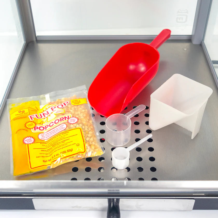 A bag of Fun Pop popcorn kernels, a red plastic scoop, and measuring cups sit on the metal surface of a countertop popcorn machine with holes, a glass enclosure, and a stainless steel kettle.