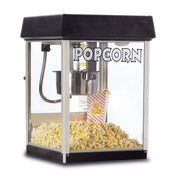 A countertop popcorn machine with a black top and base, transparent glass sides, and the word POPCORN in bold white letters. Inside, fresh popcorn surrounds a stainless steel kettle, with a striped popcorn bag labeled POP CORN filled to the brim.