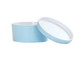 A round, light blue Bento Box with a matching, slightly raised lid leaning against it. Made from melamine, both the container and glossy lid feature white interiors and are dishwasher safe. The background is plain white.