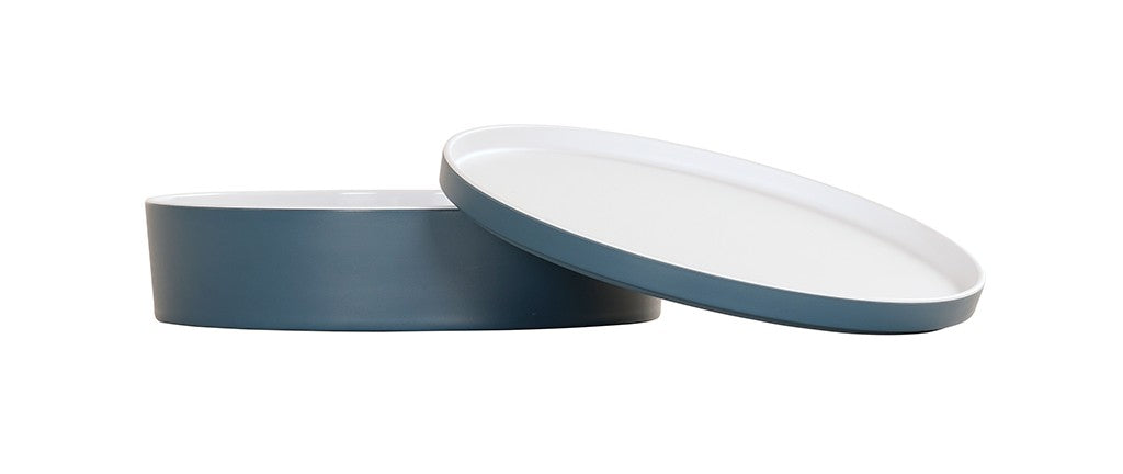 A round, two-piece bento box with a white melamine interior and blue exterior. The cylindrical base and angled lid reveal both top and inside surfaces. Dishwasher safe, it sits against a plain white background.