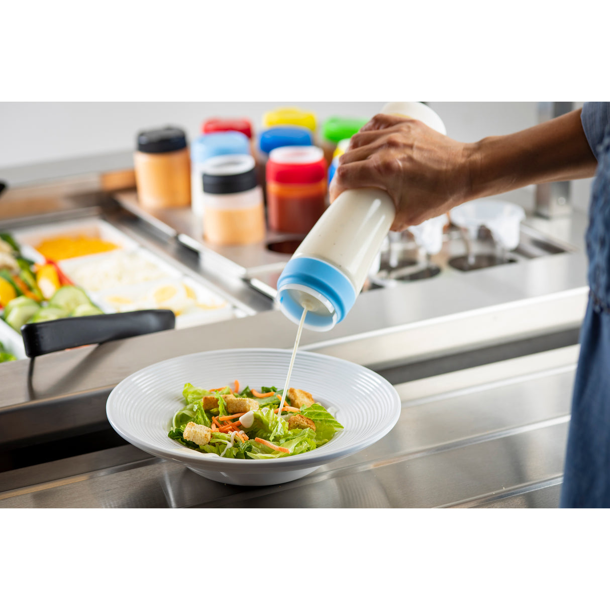 A person pours creamy dressing from a ValveTop squeeze dispenser onto a Caesar salad with lettuce and croutons in a white bowl. In the background, colorful NSF-certified condiment bottles and various salad ingredients are visible on a stainless steel counter.
