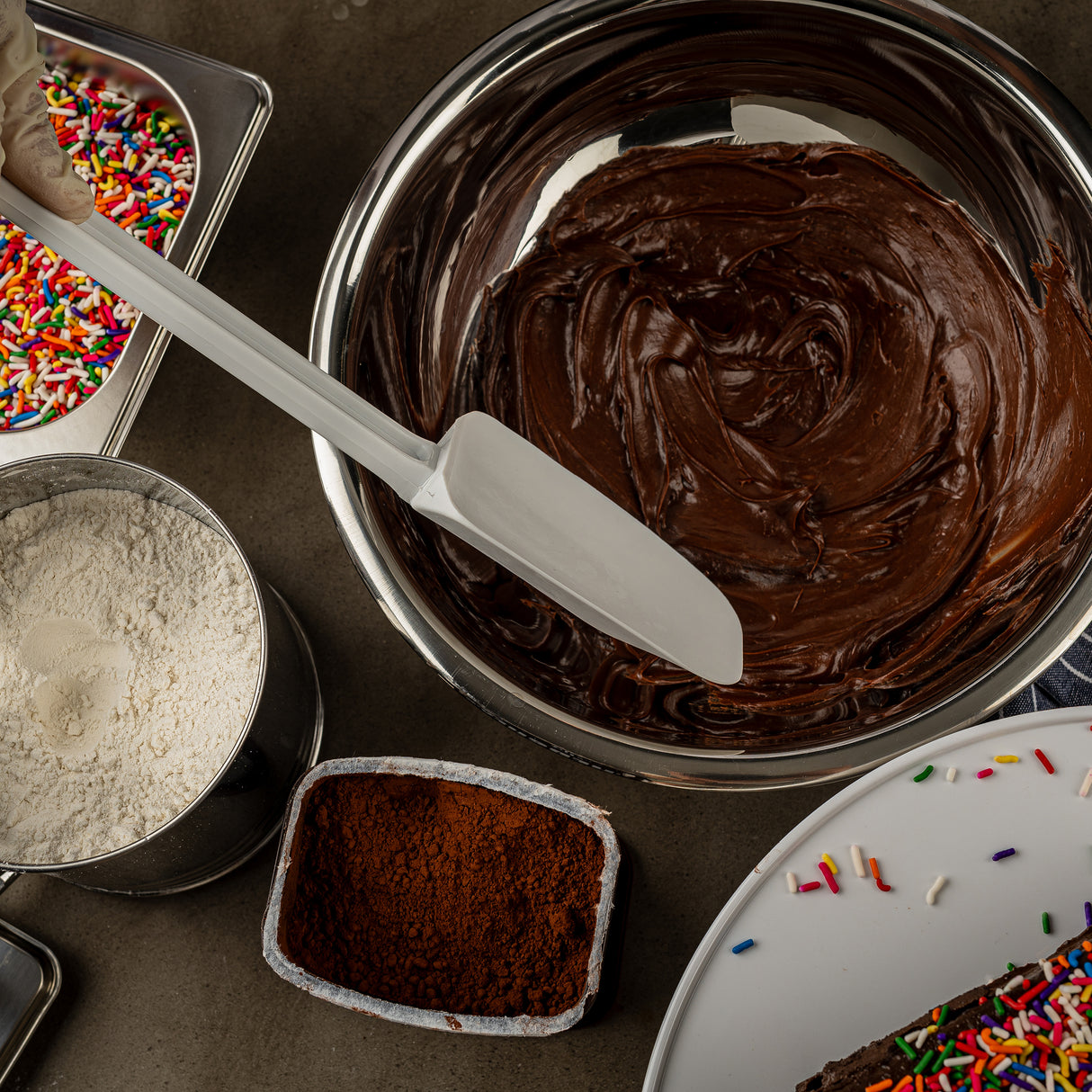 A metal bowl filled with smooth chocolate cake batter is being mixed with a heat resistant spatula. Nearby are bowls of flour, cocoa powder, and rainbow sprinkles on a dark countertop, with a decorated cake partially visible in the corner.