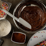 A metal bowl filled with smooth chocolate cake batter is being mixed with a heat resistant spatula. Nearby are bowls of flour, cocoa powder, and rainbow sprinkles on a dark countertop, with a decorated cake partially visible in the corner.
