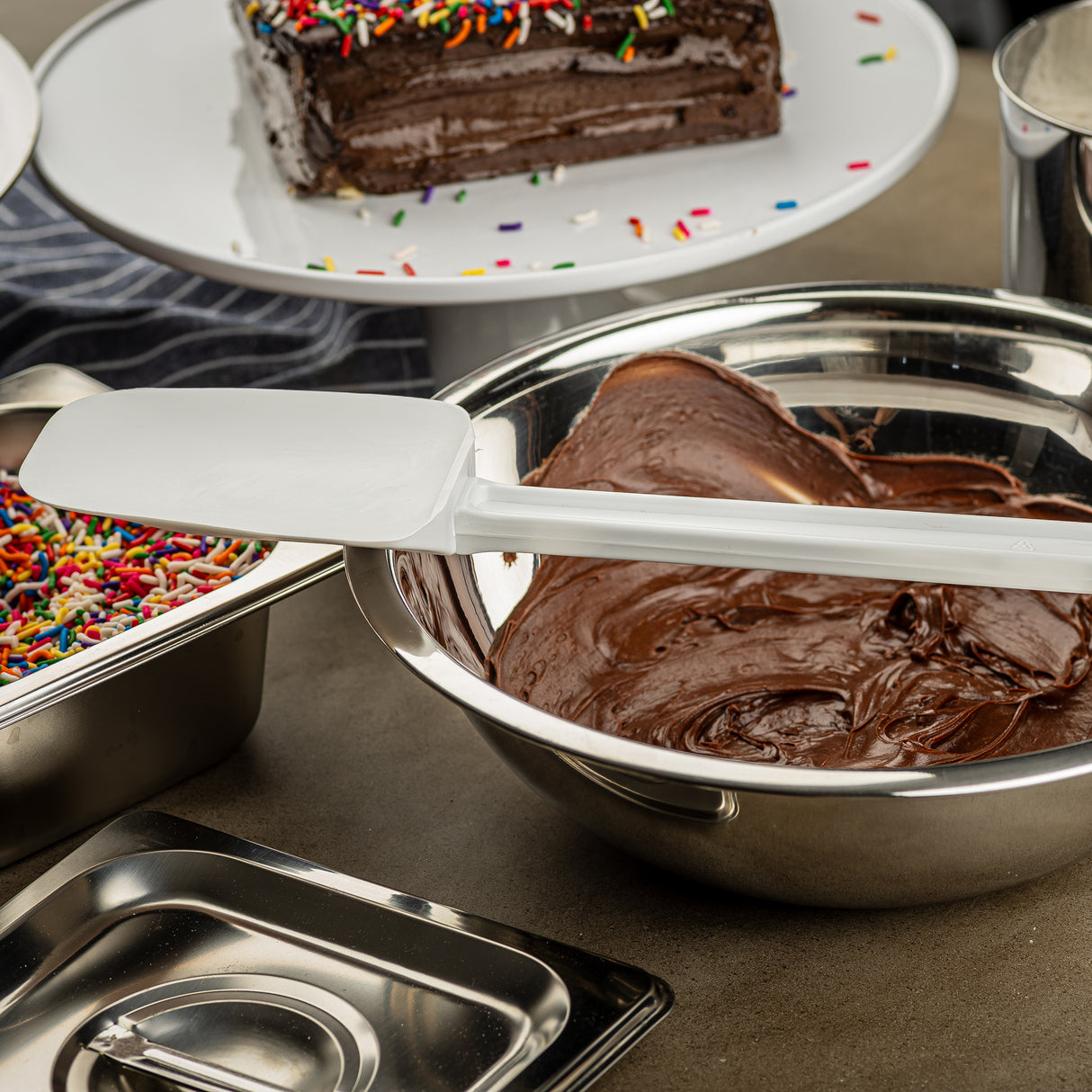 A metal bowl filled with chocolate frosting and a BPA free spatula sits on a counter. Nearby are a tray of colorful sprinkles and a metal lid. In the background, a chocolate cake with rainbow sprinkles rests on a white platter.