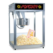 A metal E-Z Kleen kettle popcorn machine with a glass enclosure is filled with popcorn. Inside, a red and yellow popcorn box sits on the pile. The machine’s top features a POPCORN sign in bright retro-style letters.