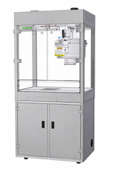 A metal industrial machine with a transparent front panel, control buttons on the top left, and a stainless steel cabinet below. Inside, the Discovery 32 oz. Popcorn Popper features a robotic arm mechanism and visible warning labels for safety.