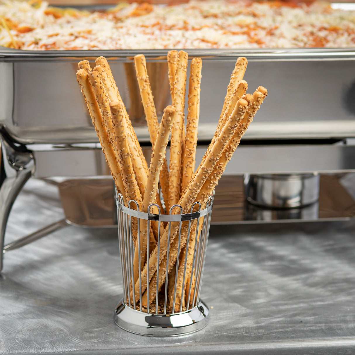 A TableCraft stainless steel bread stick holder displays several long, thin, sesame-covered breadsticks standing upright on a silver table. In the background, a large chafing dish is filled with baked pasta topped with melted cheese.