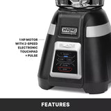 Close-up of a black Waring Commercial bar blender base showing buttons labeled Low, High, Pulse, and Stop on a touchpad. Text beside it reads 1 HP Motor with 2-speed electronic touchpad + pulse. Features copolyester container at the bottom.