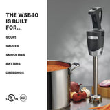 A black handheld immersion blender stands upright beside a stainless steel pot filled with tomato soup. Chopped onions, tomatoes, garlic, and peppers are on the counter. Text reads: The medium duty WSB40 handles soups, sauces, smoothies—up to 24 qt capacity. Certification logos below.