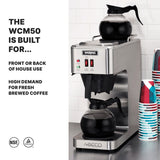 A stainless steel pourover coffee brewer with self regulating warmers holds two glass carafes, one on top and one at the base. Disposable cups are stacked beside it. Text reads: “THE WCM50 IS BUILT FOR… FRONT OR BACK OF HOUSE USE, HIGH DEMAND FOR FRESH BREWED COFFEE.”.