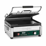 A stainless steel panini grill with a ridged top plate open at an angle and a flat bottom plate. It features a black handle, indicator lights, temperature control knob, and the “Waring Toastato Supremo” label on the front panel.
