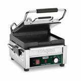 A compact toasting grill, this stainless steel Waring panini press features flat cast iron plates, a black handle, and a partially open top plate. The front panel offers adjustable thermostats, indicator lights, and the engraved Waring brand name.