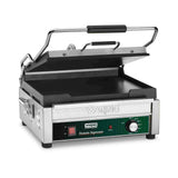 A stainless steel Waring panini press with a black handle and partially open lid. The front features a green temperature control dial, a red power switch, indicator lights, and “Tostato Supremo” text. This large toasting grill has flat cast iron plates.