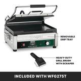 A stainless steel Panini Grill with a black grill plate and handle is shown partly open. Text points to a removable drip tray and a heavy-duty grill brush with scraper, both included with the WFG275T model.