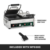 A stainless steel Waring commercial panini press featuring cast iron plates, a raised top plate, and labeled features: removable drip tray and heavy-duty grill brush with scraper—both included with model WFG300. Black control knobs and indicator lights are visible on the front panel.