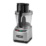A silver Waring food processor with a clear 4 qt batch bowl and black handle. The base features three buttons: green, red, and black, with icons and text. The lid has a feed tube, pusher, and LiquiLock seal system. Waring logo on the front.