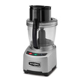 A heavy duty silver Waring food processor with a clear plastic bowl, black handle, and black lid. The base has green, red, and black control buttons and the Waring logo on the front, shown against a plain white background.