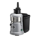 A silver and black heavy duty Juice Extractor with a cylindrical metal body, vertical black feed chute, side spout, and large black pulp container on the right. “Waring” is on the front, with a green power switch and continuous pulp ejection system.