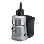 A silver and black heavy duty Juice Extractor with a cylindrical metal body, vertical black feed chute, side spout, and large black pulp container on the right. “Waring” is on the front, with a green power switch and continuous pulp ejection system.