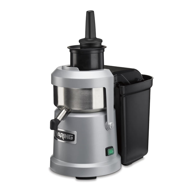 A silver and black heavy duty Juice Extractor with a cylindrical metal body, vertical black feed chute, side spout, and large black pulp container on the right. “Waring” is on the front, with a green power switch and continuous pulp ejection system.