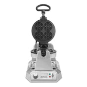 A stainless steel heavy duty waffle maker with its lid open reveals circular, four-sectioned non-stick waffle plates. The control panel at the base has a dial knob, two indicator lights (green and red), and the brand name “Waring.”.