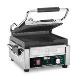 A stainless steel Waring commercial panini grill with a black handle, ribbed cast iron plates, and a control panel featuring a power switch, ready light, temperature dial, and green and red indicator lights. The lid is partially open above the grill plates.