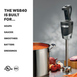 A black immersion blender stands upright near a pot of tomato soup, surrounded by onions, garlic, tomatoes, and a red pepper. Perfect for soups and sauces, this medium duty model handles up to 24 qt. capacity. Certification logos are at the bottom.
