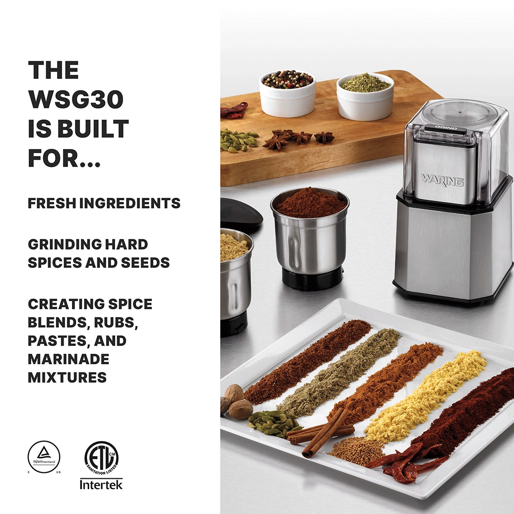 A professional spice grinder sits on a counter next to bowls of spices, three stainless steel grinding cups, and a tray with colorful ground spices and whole dried chilies. Text on the left lists uses: grinding fresh ingredients, hard spices, seeds, and making blends.