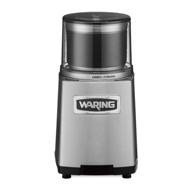 A stainless steel Waring WSG60KSDI Commercial Spice Grinder with a clear plastic top, labeled “Waring” on the front. The brushed metal finish and lock/unlock symbols beneath the transparent lid make it ideal for international use.