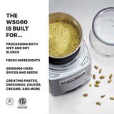 A silver Commercial Spice Grinder filled with greenish-yellow powder sits on a white surface beside its clear lid and scattered cardamom pods. Text on the left reads: "The Waring WSG60E is built for international use, processing both wet and dry blends, and more.