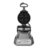 A stainless steel Belgian Waffle Maker with a round, open black waffle plate showing a grid pattern sits on a square base. The front panel has a handle, control knobs, and indicator lights. The hinged lid stands upright, revealing the interior.