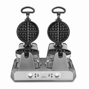 A double commercial waffle maker with two open round, non-stick plates, black handles, and a silver base. Two control knobs, indicator lights, and warning labels are on the front. The machine is positioned on a plain white background.