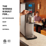 A hand dispenses hot water from a tall, stainless steel 5 gallon Waring Hot Water Dispenser into a mug. The digital display shows 205°F. This countertop unit is ideal for hot beverages, soup, grits, and oatmeal. Two people sit at a booth in the background.