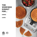 A heavy-duty waffle maker sits on a white surface with a round waffle inside. Three golden waffles, some sliced, and a bowl of fresh strawberries are nearby. Text reads: THE WWD180X IS BUILT FOR... DINERS • CAFÉS. Certification logos are below.