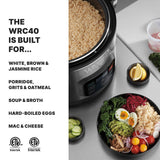 A commercial rice cooker filled with cooked rice sits on a counter next to bowls of fresh food: cucumber, hard-boiled eggs, mixed greens, shredded carrots, red cabbage, beetroot, and avocado. Text lists foods the PFAS-free WRC40 cooker can make.