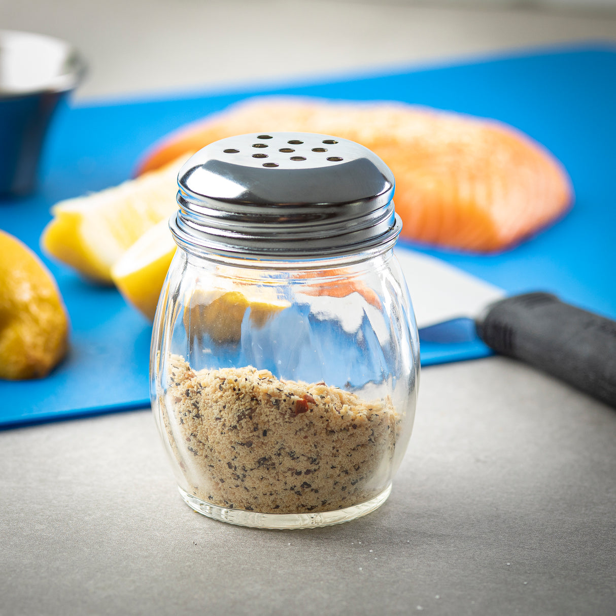 A BPA free glass spice shaker with a metal lid sits on a gray surface. Inside, mixed seasoning with black and tan specks is visible. In the blurred background are lemon wedges, salmon fillet, a blue cutting board, and a metal spatula.