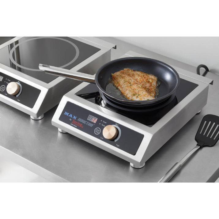 A breaded fish fillet cooks in a black frying pan on a stainless steel commercial induction cooker. The digital display shows “Max.” A countertop range, spatula, and metal surfaces add to the clean, modern kitchen setting.