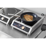 A breaded fish fillet cooks in a black frying pan on a stainless steel commercial induction cooker. The digital display shows “Max.” A countertop range, spatula, and metal surfaces add to the clean, modern kitchen setting.