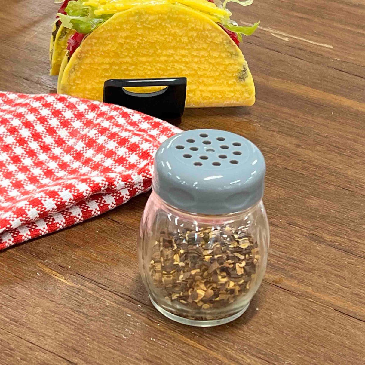 A small glass cheese shaker with a gray lid, similar to the classic TableCraft Products Swirl Cheese Shaker, is filled with crushed red pepper flakes and sits on a wooden table near a checkered cloth and a taco topped with lettuce and cheese.