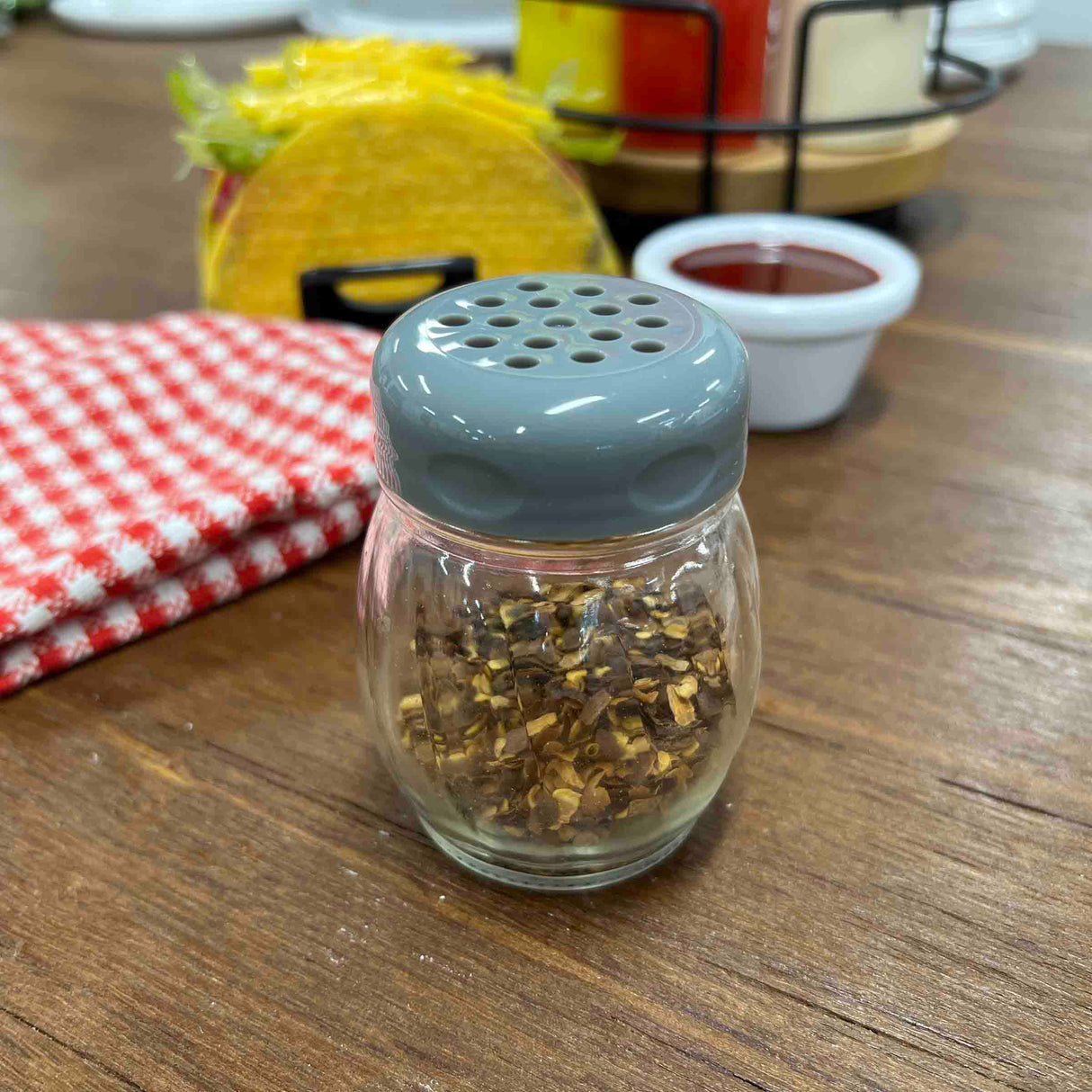 A glass cheese shaker with a gray lid, filled with crushed red pepper flakes, sits on a wooden table. Behind it are a red checkered napkin, a taco, sauce cup, and a wire rack of TableCraft Products condiment bottles.