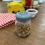 A glass cheese shaker with a gray lid, filled with crushed red pepper flakes, sits on a wooden table. Behind it are a red checkered napkin, a taco, sauce cup, and a wire rack of TableCraft Products condiment bottles.