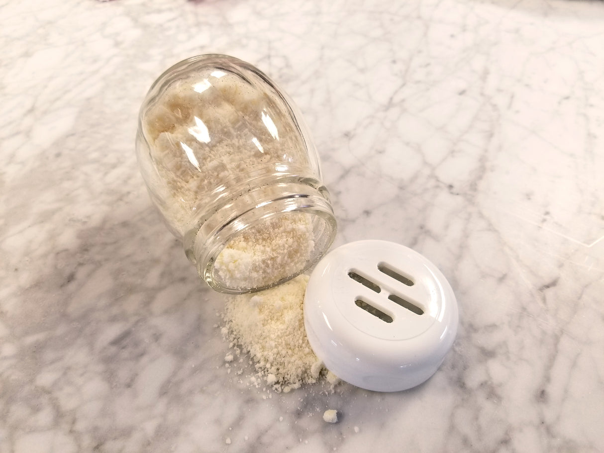 A TableCraft 6 oz glass shaker lies tipped over on a white marble surface, spilling grated parmesan cheese onto the counter. The white plastic lid rests nearby, making it easy to refill or swap with a replacement jar.