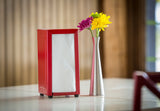 A red napkin holder with white napkins stands on a table next to a sleek zinc alloy Flower Vase with a brushed finish, holding three colorful flowers—yellow, purple, and magenta—against a softly blurred background.