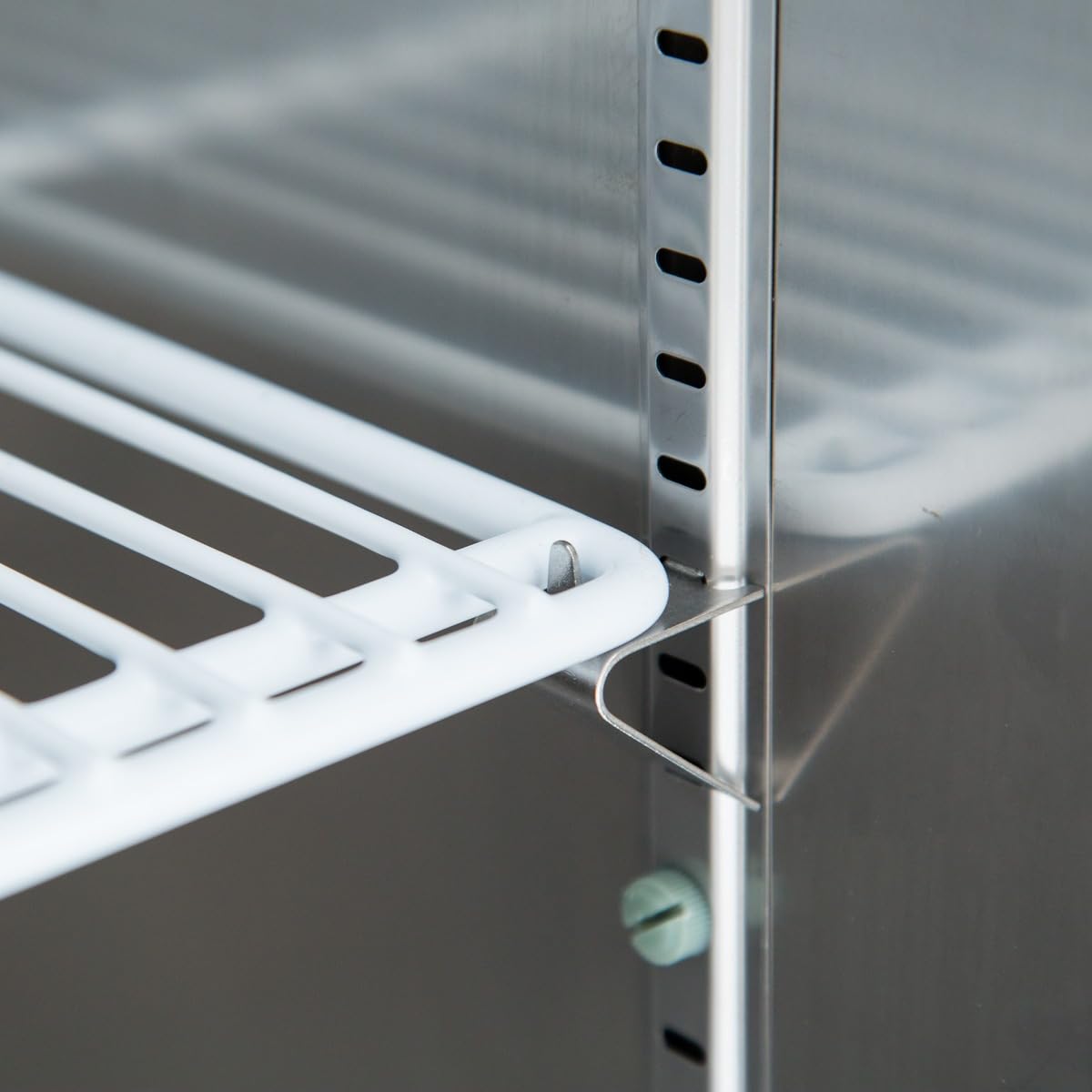 Close-up of a white wire shelf installed on a metallic shelving unit inside a commercial refrigerator. The shelf is supported by a metal bracket slotted into vertical rails, which feature evenly spaced holes for adjustable placement.